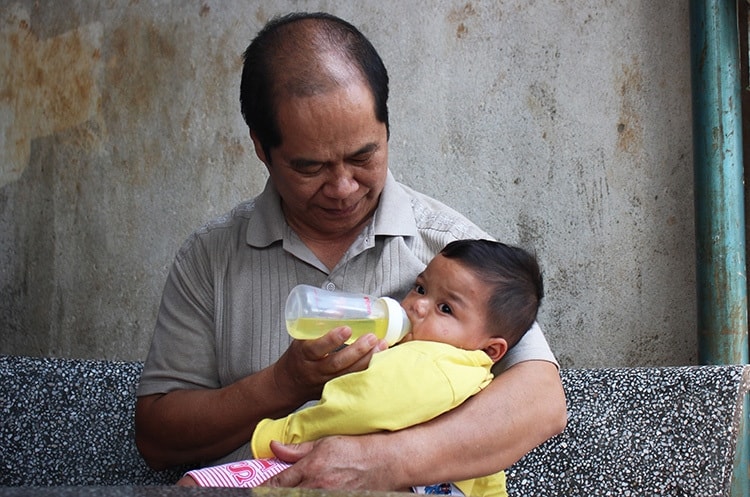 Ông Nhật và bé Đinh Thiên Đức, 11 tháng tuổi. Vừa lọt lòng, bé Đức đã bị bỏ rơi trên rẫy và được ông Nhật mang về nuôi. Bé bị phát hiện bệnh tim bẩm sinh từ khi 2 tháng tuổi, cha nuôi đã phải bán bò để đưa bé đi mổ. Sau hai lần phẫu thuật, đến nay bé Đức đã ổn. Ảnh: Phan Diệp.