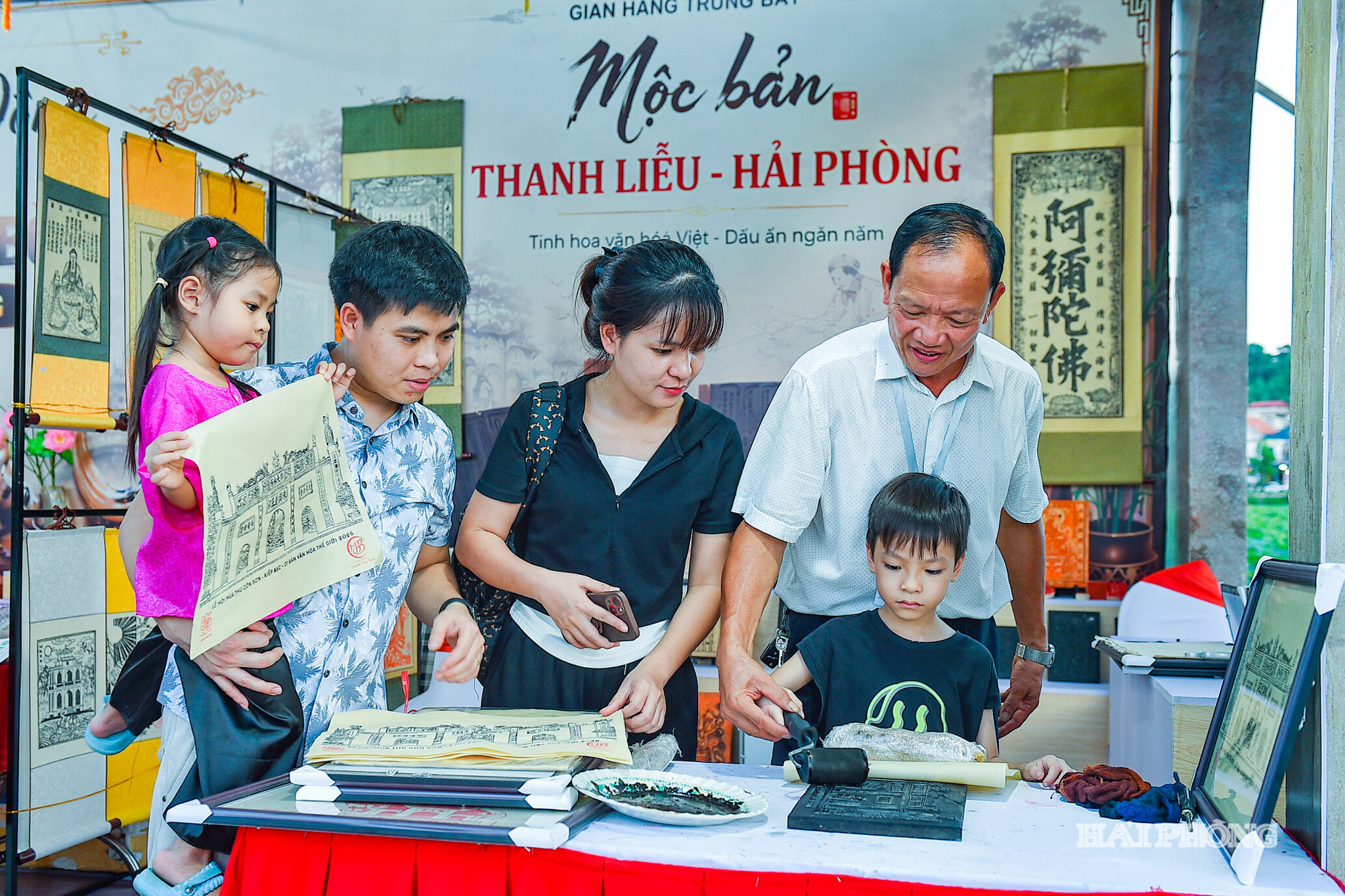 Nghệ nhân hướng dẫn một em nhỏ trải nghiệm kỹ thuật in mộc bản Thanh Liễu tại Tuần Văn hóa, Du lịch và Xúc tiến thương mại. Hoạt động giúp thế hệ trẻ trực tiếp khám phá nghề truyền thống, qua đó thêm hiểu và yêu những giá trị văn hóa được gìn giữ qua nhiều thế hệ.